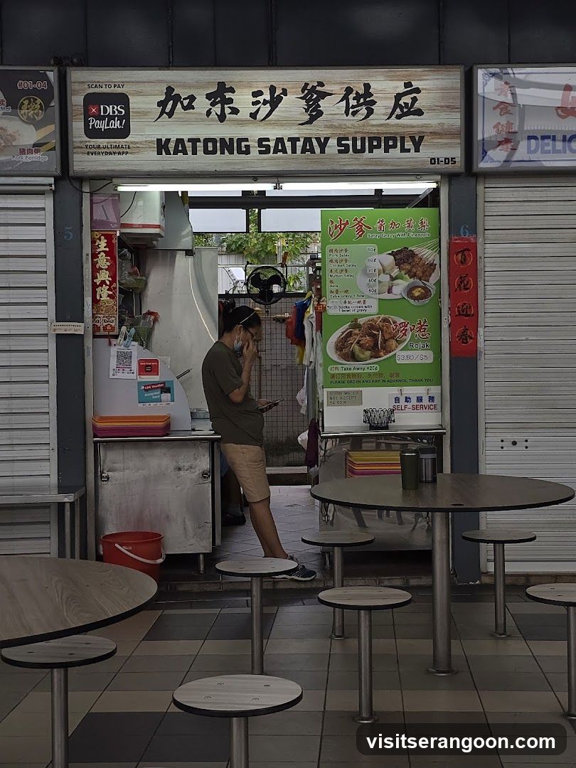 A storefront with a sign that reads "KATONG SATAY SUPPLY" in English and Chinese characters. There is a menu board to the right of the entrance displaying satay and rojak. Tables and stools are set up in front of the shop.