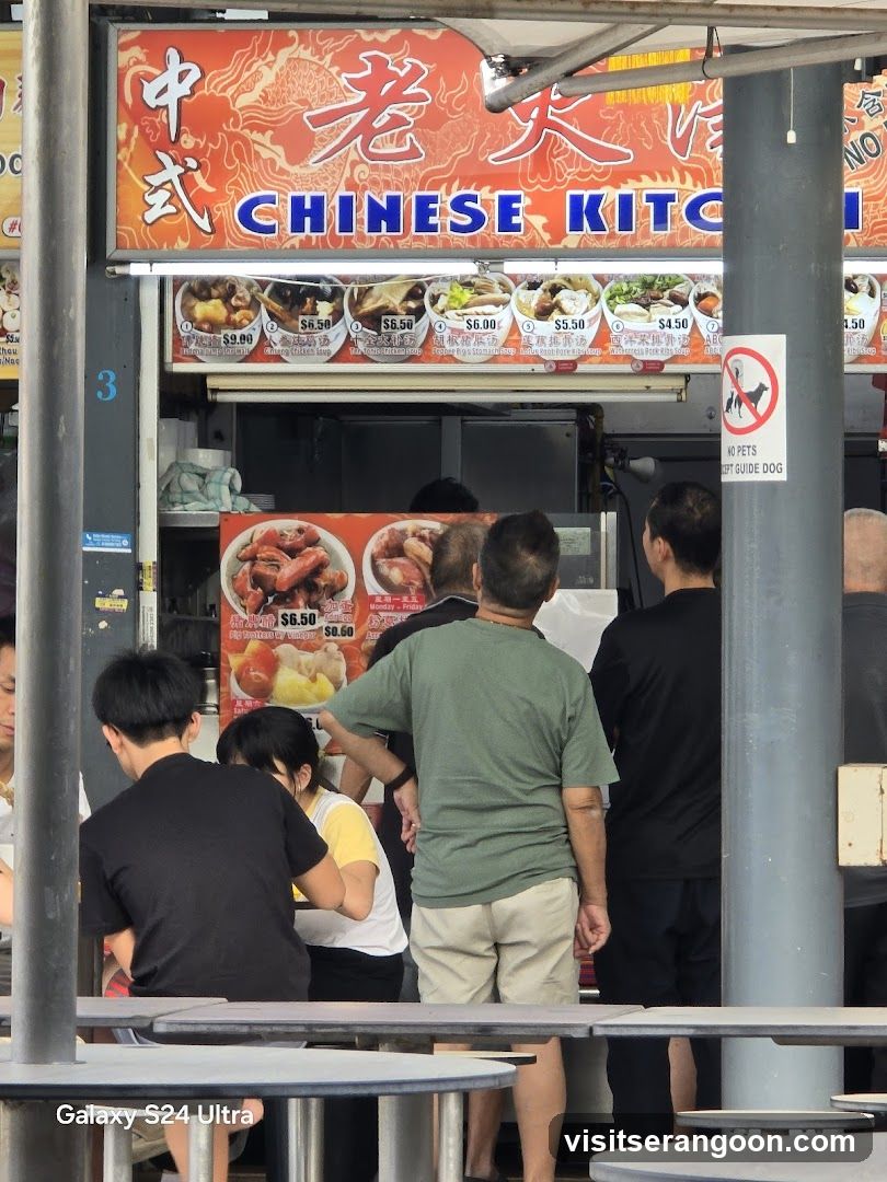 A busy outdoor food stall with a prominent sign reading "CHINESE KITCHEN" in English and Chinese characters. Below the main sign, a menu displays various soup dishes with prices. Several people are queuing or dining at tables in front of the stall. A "No Pets Except Guide Dog" sign is visible on the right.