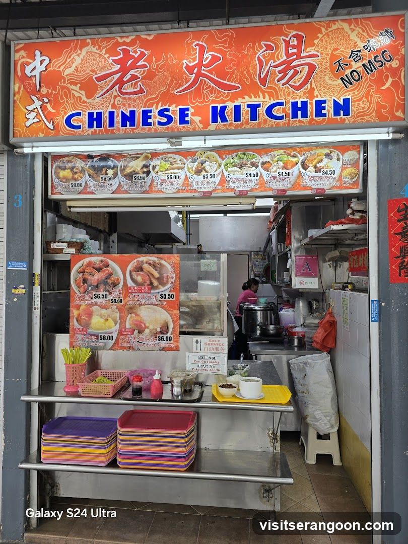A food stall with a large orange sign that says "CHINESE KITCHEN" in English and Chinese characters. Below the sign are pictures of various soups and dishes with prices. The stall appears to be open, with food preparation visible in the background.