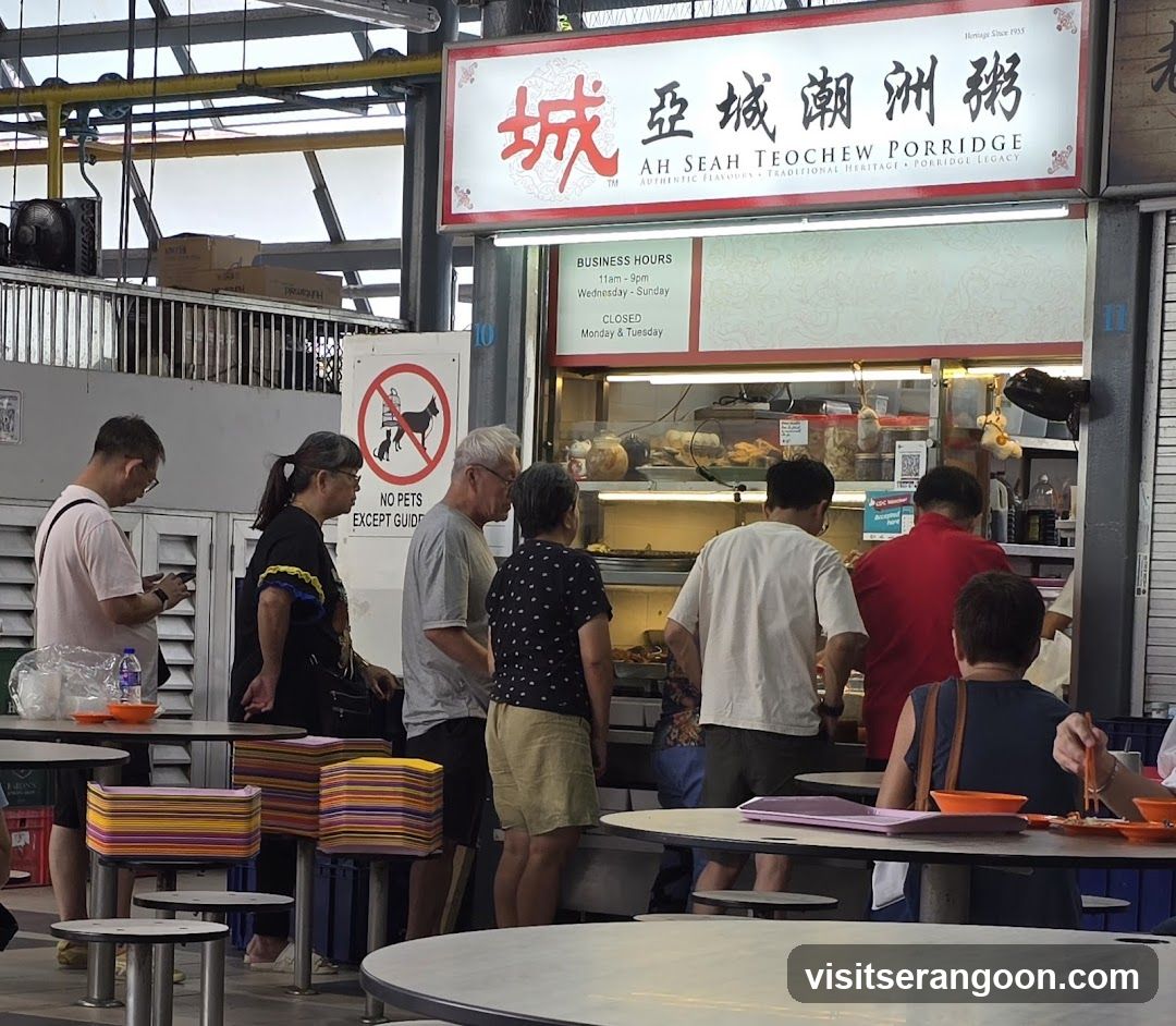 A food stall with a sign that reads "Ah Seah Teochew Porridge" and lists business hours. Several people are queuing and being served. There is also a sign prohibiting pets.