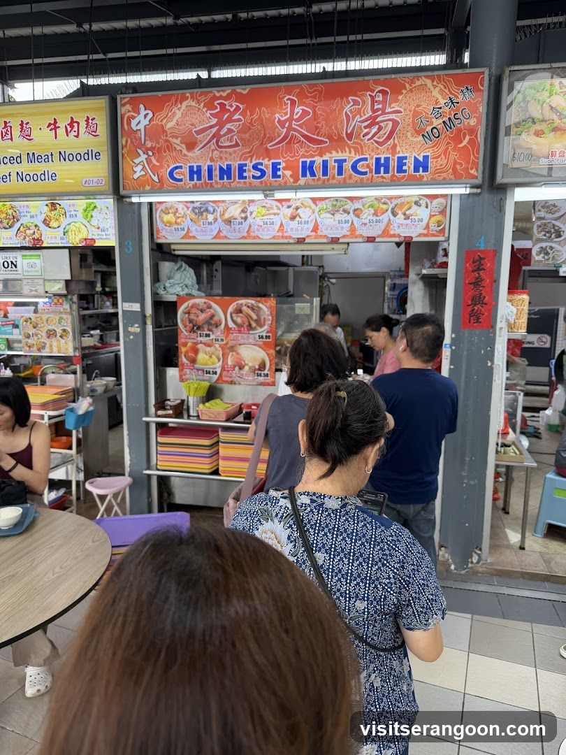 A busy food stall with a prominent orange sign displaying "CHINESE KITCHEN" in blue letters and Chinese characters above. Below the main sign, a menu board shows various dishes with prices. Several people are queuing to order. The stall appears to be part of an outdoor food court or market.