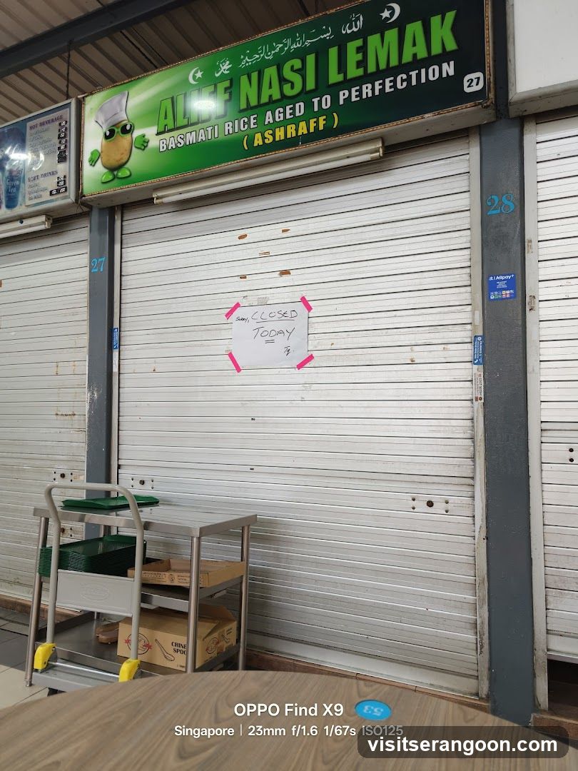 The storefront of Aliff Nasi Lemak, a food stall, is shown with its metal shutters down. A sign above the shutters reads "ALIFF NASI LEMAK BASMATI RICE AGED TO PERFECTION (ASHRAFF)". A handwritten sign on the shutter says "Sorry, CLOSED TODAY". A trolley with green containers and boxes is visible to the left.