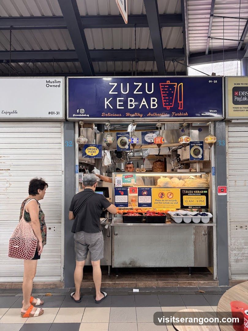 A Muslim-owned kebab stall with a blue sign that reads "ZUZU KEBAB" and lists "Delicious. Healthy. Authentic Turkish." Below the sign, there is a counter with food items and a staff member serving customers. Signs indicate "Order Here" and "Collect Here." The operating hours are "Open Daily 10 AM - 9 PM."