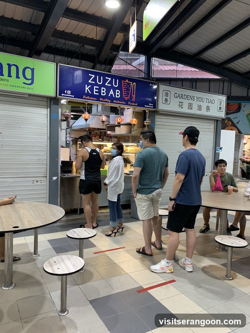 A food stall with a blue sign that reads "ZUZU KEBAB" and lists "Delicious. Healthy. Authentic. Turkish." Below the sign, there are colorful hanging lamps and people queuing to order. To the right of the kebab stall is another stall with a sign that reads "GARDENS YOU TIAO" in English and Chinese.