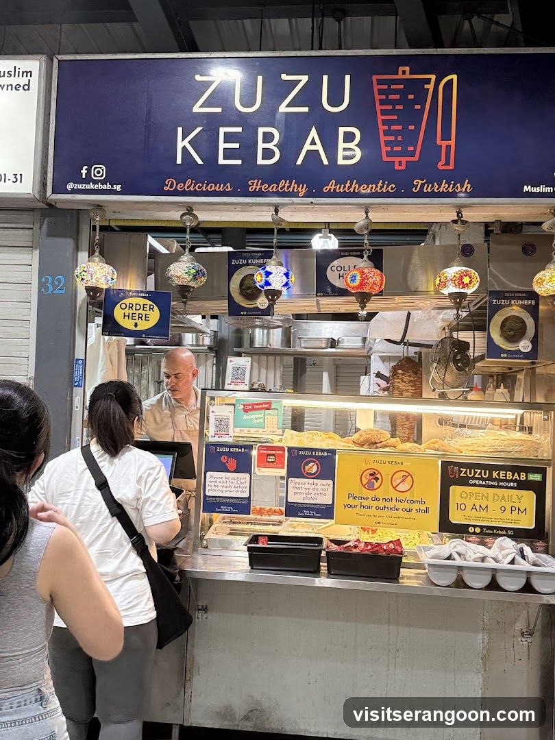 A food stall with a large sign that says ZUZU KEBAB. Below the sign are decorative lamps and a display case with food. There are also signs with operating hours and rules.