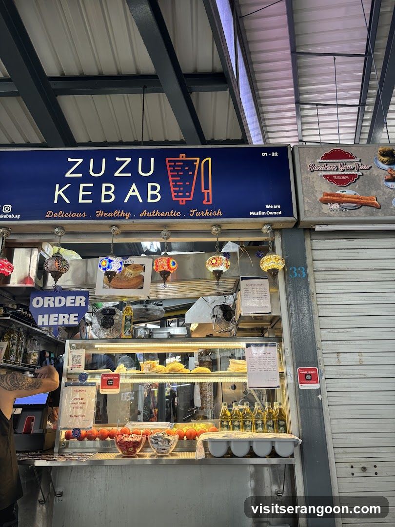 A food stall with a prominent blue sign displaying "ZUZU KEBAB" and the tagline "Delicious. Healthy. Authentic. Turkish.". Below the sign, there is a display counter with food items, bottles of olive oil, and decorative lamps. An "ORDER HERE" sign is visible on the left.
