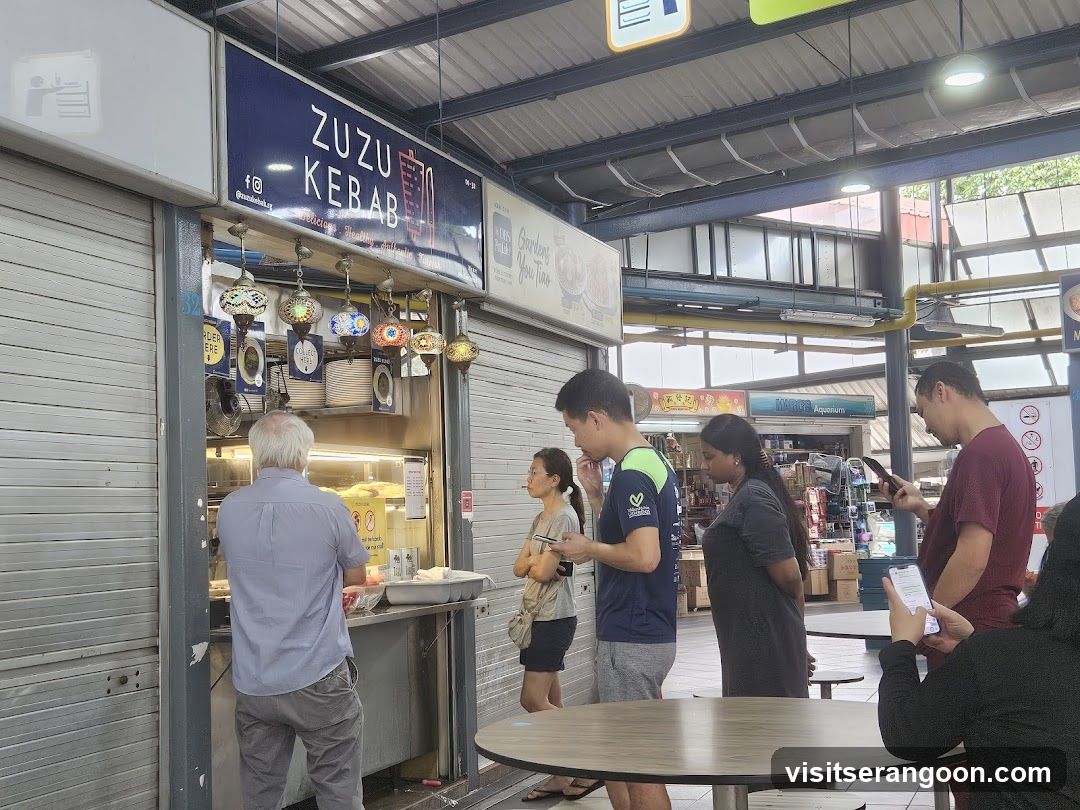 A food stall with a blue sign that says "ZUZU KEBAB" and lists "Delicious, Healthy, Authentic Turkish". There are several decorative lamps hanging above the counter. People are standing in line to order.