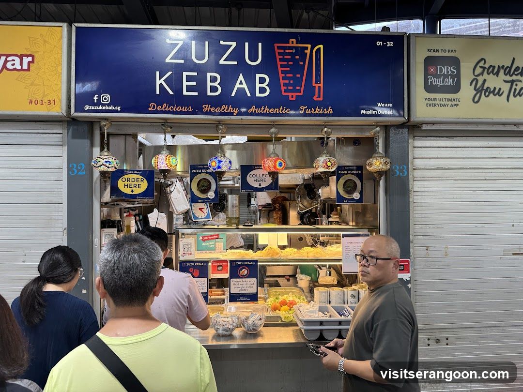 A food stall with a blue sign that reads "ZUZU KEBAB" and lists "Delicious. Healthy. Authentic. Turkish." Below the sign are three decorative lamps and various signs, including one that says "ORDER HERE." People are standing in front of the stall, and food is visible behind the counter.