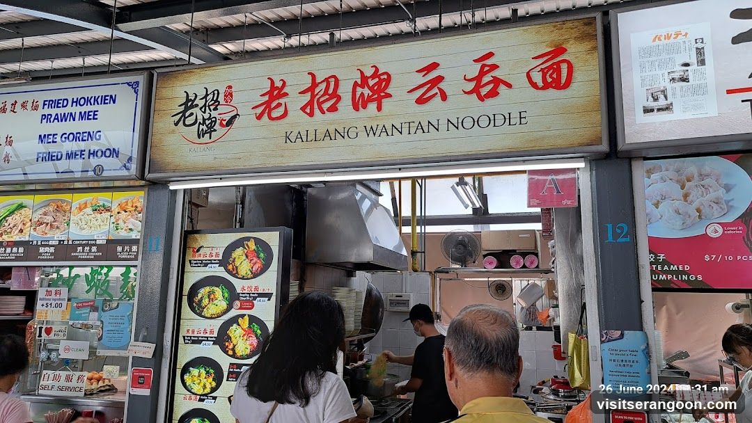 A food stall with a large sign displaying "KALLANG WANTAN NOODLE" in English and Chinese. Below the main sign, there are smaller signs with pictures and descriptions of various noodle dishes and fried Hokkien prawn mee. To the right, a sign advertises steamed dumplings. People are seen ordering and staff are preparing food in the background.