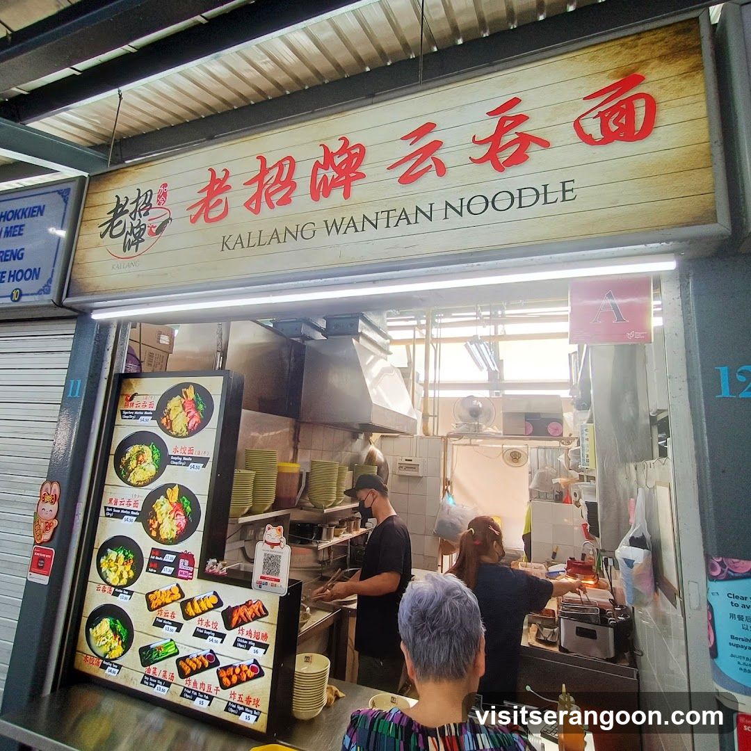A food stall with a prominent sign reading "KALLANG WANTAN NOODLE" in English and Chinese characters. Below the sign, a menu board displays various noodle dishes and fried items with prices. Two staff members are visible working in the kitchen area behind the counter.