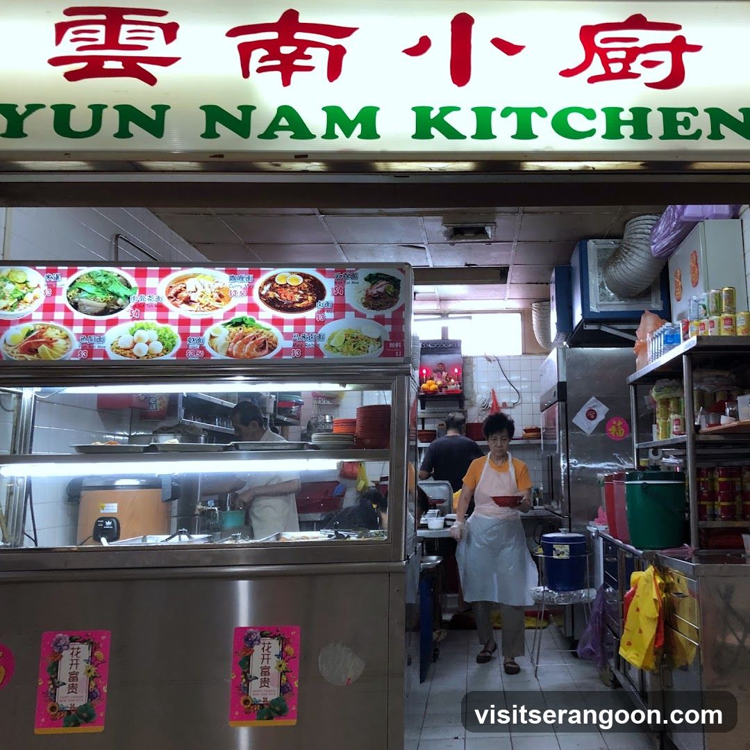 A food stall with a sign that reads "Yun Nam Kitchen" in English and Chinese characters. Below the sign is a menu board displaying various noodle dishes with prices. Inside the stall, staff members are preparing food.
