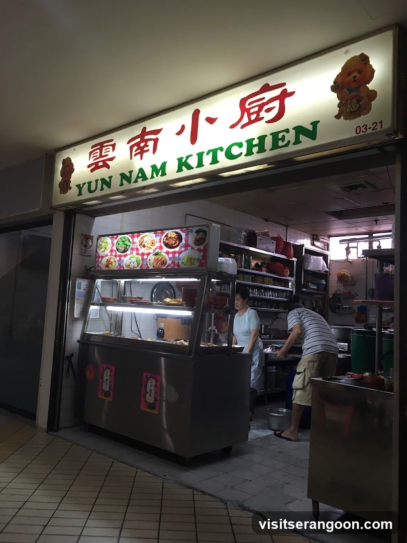A brightly lit storefront with a large illuminated sign that reads "YUN NAM KITCHEN" in English and Chinese characters. The sign also features a cartoon bear graphic. Below the sign, a display case showcases various food items. Two staff members are visible inside the establishment.