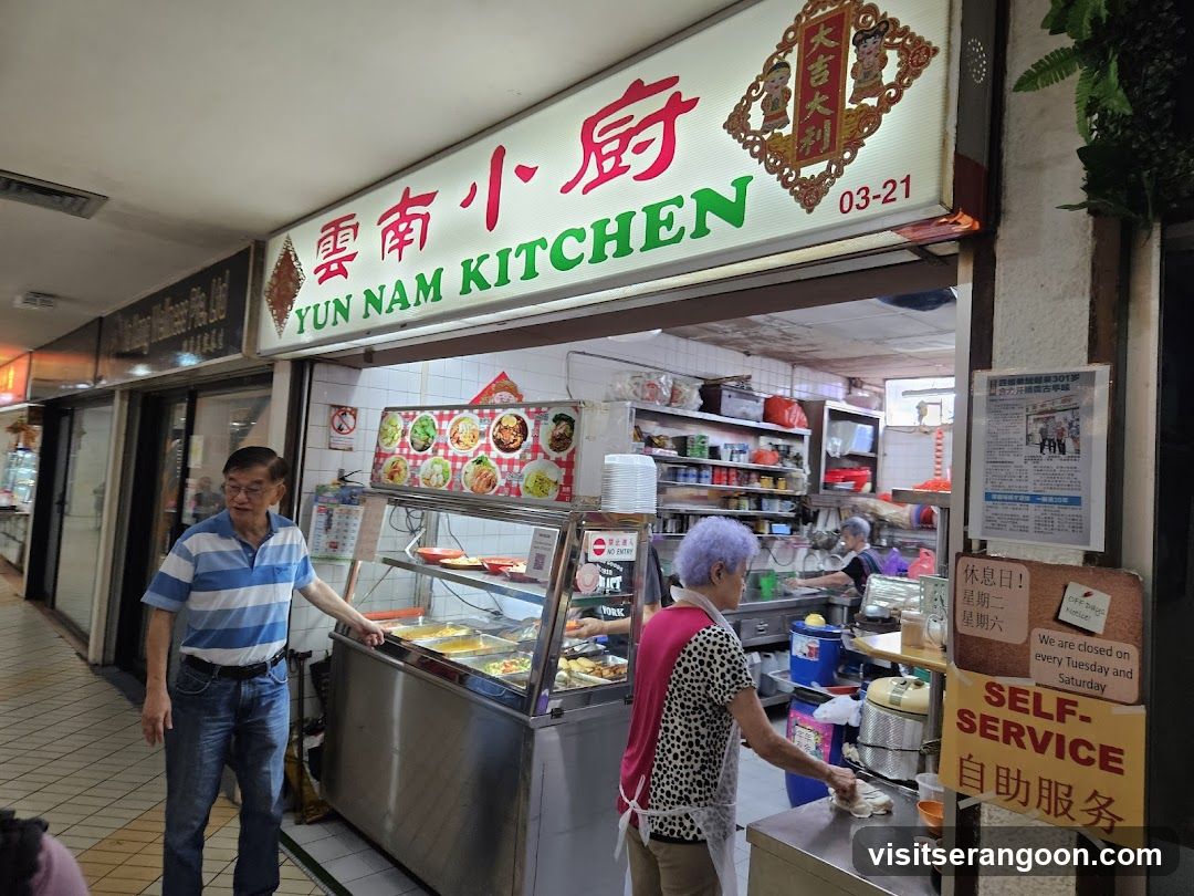 The exterior of a food stall with a prominent sign reading "YUN NAM KITCHEN" in English and Chinese. A man in a blue and white striped shirt stands to the left, gesturing towards a display of food. An older woman with purple hair is seen cleaning a counter to the right. Shelves stocked with various items are visible behind the counter. A sign indicates that the establishment is closed on Tuesdays and Saturdays and offers self-service.