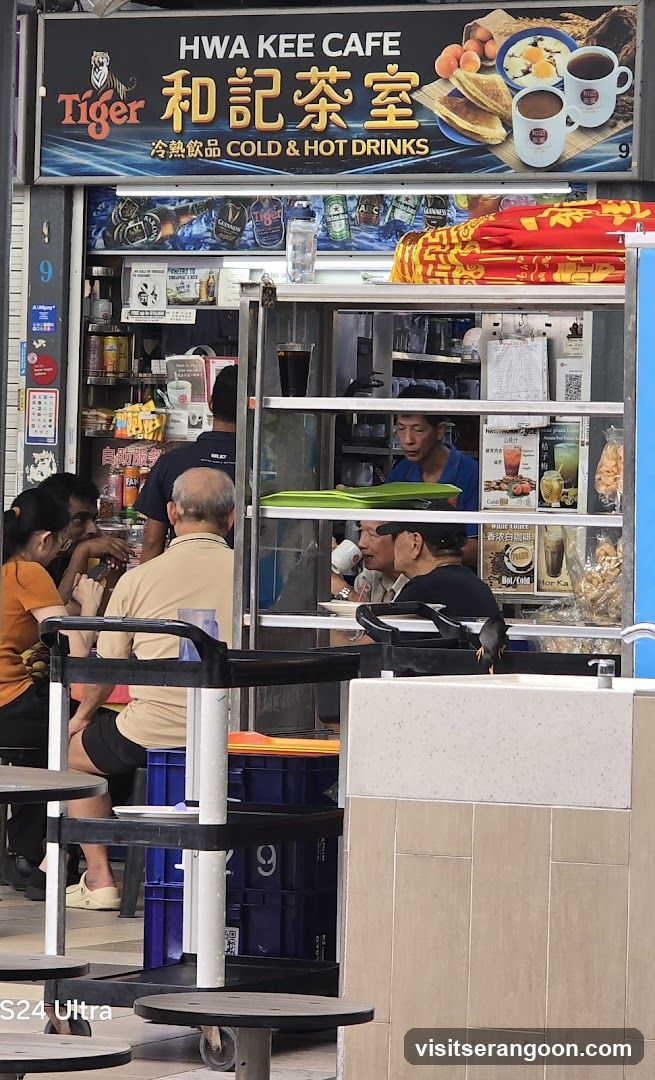 The exterior of a cafe with a prominent sign displaying "HWA KEE CAFE" in English and Chinese characters, along with "Tiger" branding and "COLD & HOT DRINKS". The sign also features images of food and drinks. Below the main sign, shelves are visible with various bottles and products. People are seated at tables in the foreground and inside the cafe.