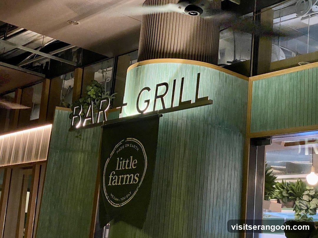A neon sign that reads "BAR + GRILL" is illuminated above a dark green banner with the text "little farms" in a circle. The banner also says "MADE ON EARTH" and "WE FARM. YOU EAT." The building facade is covered in vertical green tiles with wooden trim.