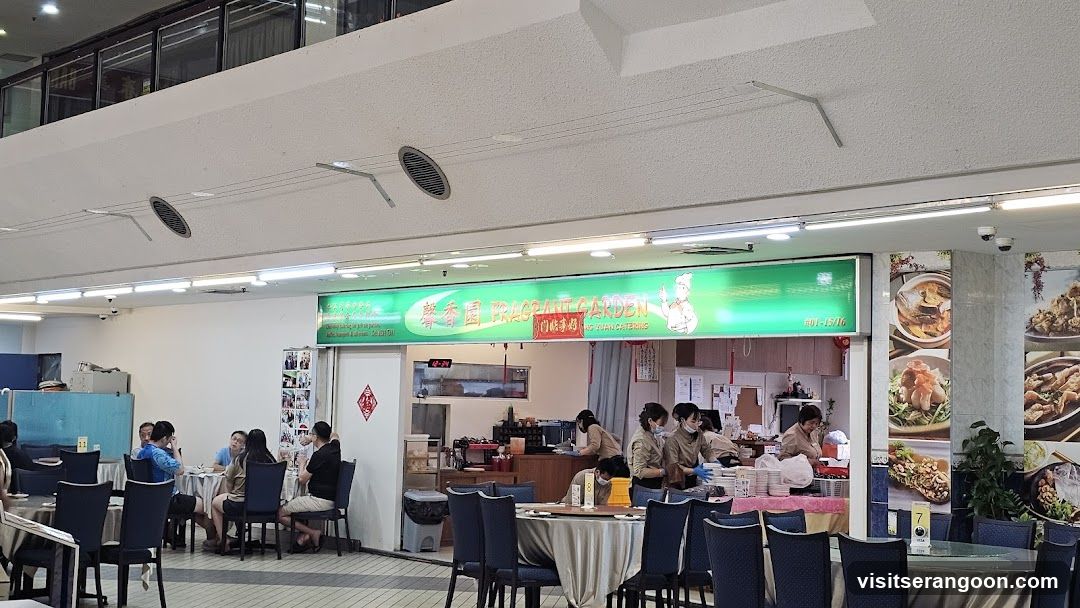 The exterior of a restaurant with a green illuminated sign that reads "Fragrant Garden" and "XUAN CATERING". There are images of food displayed on the wall to the right of the entrance. Inside, staff members are visible preparing food and serving customers. Tables are set with tablecloths and chairs.