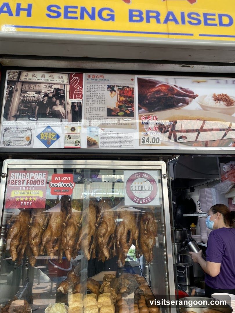 A storefront display featuring hanging braised ducks, braised duck rice, and various awards and accolades. The signage above the display reads "AH SENG BRAISED".