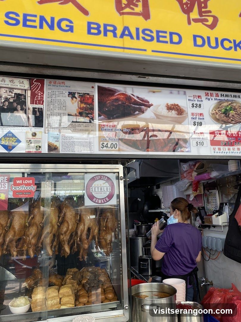 A shop front with a yellow sign that says "SENG BRAISED DUCK" in blue letters. Below the sign is a menu board with pictures and prices of braised duck dishes. To the left, there is a display case with whole braised ducks hanging and other food items. A woman in a purple shirt is working in the background.