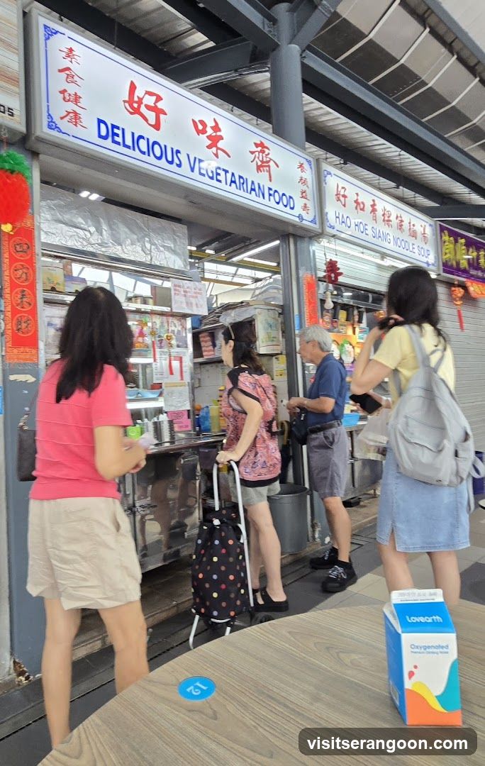 A street food stall with signage indicating "DELICIOUS VEGETARIAN FOOD" and "HAO HOE SIANG NOODLE SOUP". Several people are queuing or standing in front of the stall. A carton of Lovearth Oxygenated drink is on a table in the foreground.