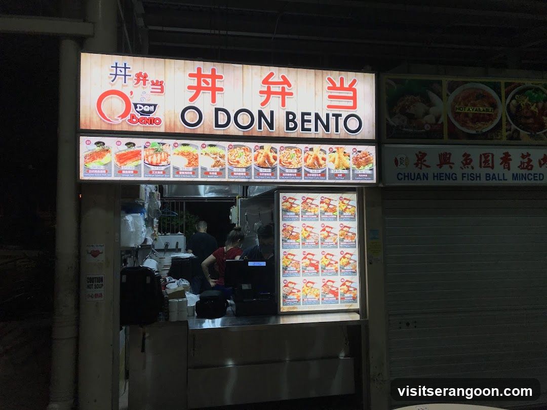 A brightly lit food stall at night, with a large sign displaying the name "O DON BENTO" in English and Chinese characters. Below the main sign, a menu board showcases various bento box meals with images and prices. To the right, another sign advertises "CHUAN HENG FISH BALL MINCED". The stall appears to be open, with staff visible inside preparing food.
