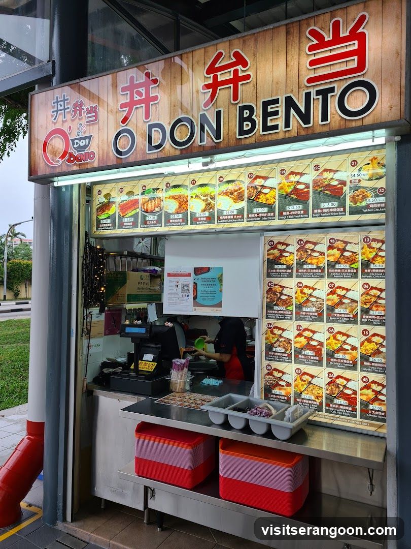 A food stall with a prominent sign displaying "O DON BENTO" in English and Japanese characters. Below the sign, a menu board showcases various bento box meals with images and prices. The stall has a counter with a cash register, stacks of colorful containers, and a person preparing food behind the counter.