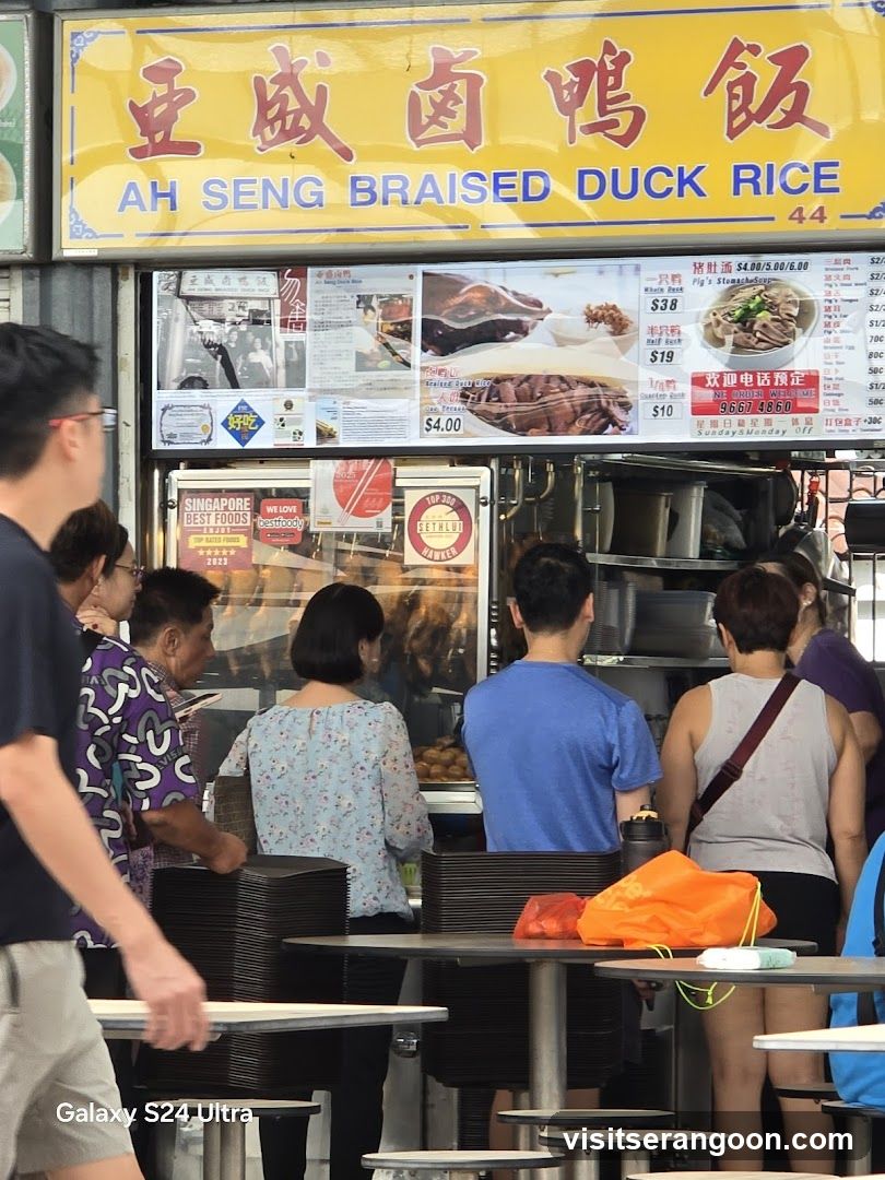 A hawker stall with a yellow sign displaying "AH SENG BRAISED DUCK RICE" in English and Chinese characters. Below the main sign, there are various menu boards and pictures of food. Several customers are queuing or standing in front of the stall. Stacks of black plates are visible on the tables.
