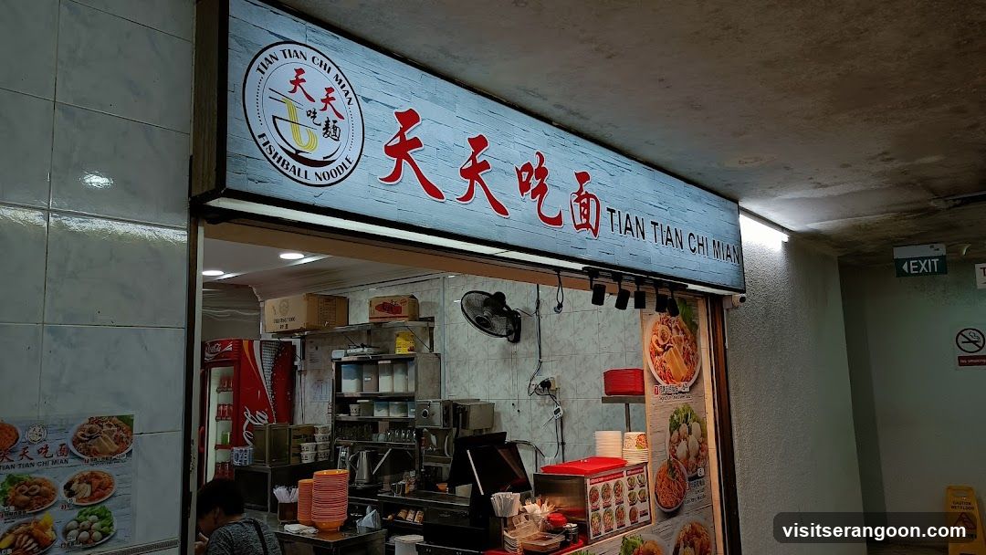 A brightly lit shop front with a large illuminated sign displaying the shop's name in both Chinese and English, "天天吃面 TIAN TIAN CHI MIAN FISHBALL NOODLE". Below the sign, the interior of the food stall is visible, showing a counter with food preparation equipment, stacks of bowls, and a Coca-Cola refrigerator. There are also posters displaying images of the food offered.