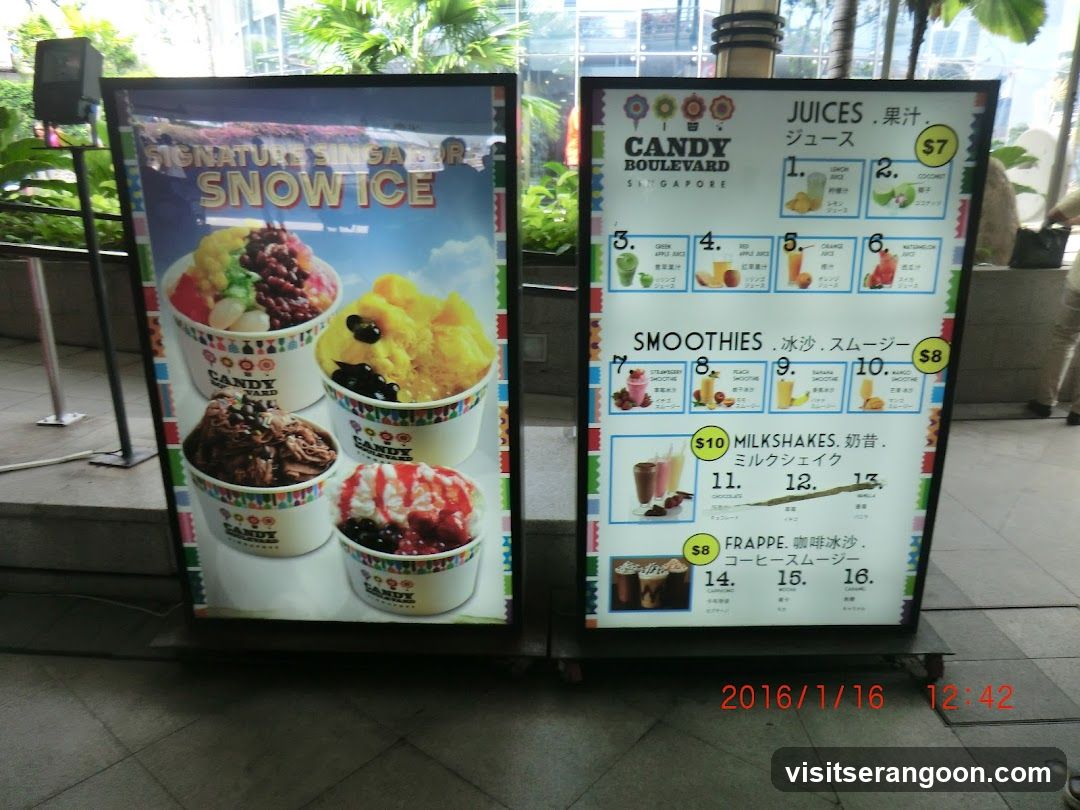 Two standing menu boards displaying drink options from Candy Boulevard Singapore. Left board features 'Signature Singapore Snow Ice' with colorful images of toppings. Right board lists juices, smoothies, milkshakes, and frappes with prices in SGD.