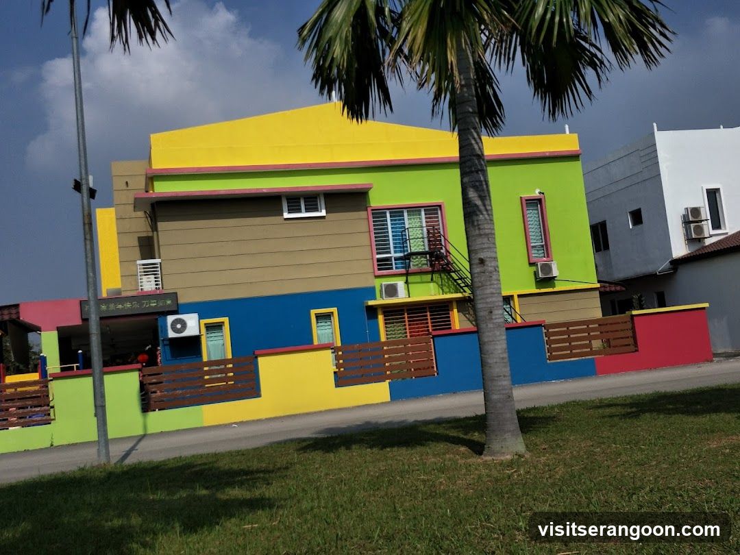 A brightly colored building with sections painted in yellow, lime green, brown, and blue. A wooden fence with horizontal slats runs along the front. Palm trees are visible in the foreground.