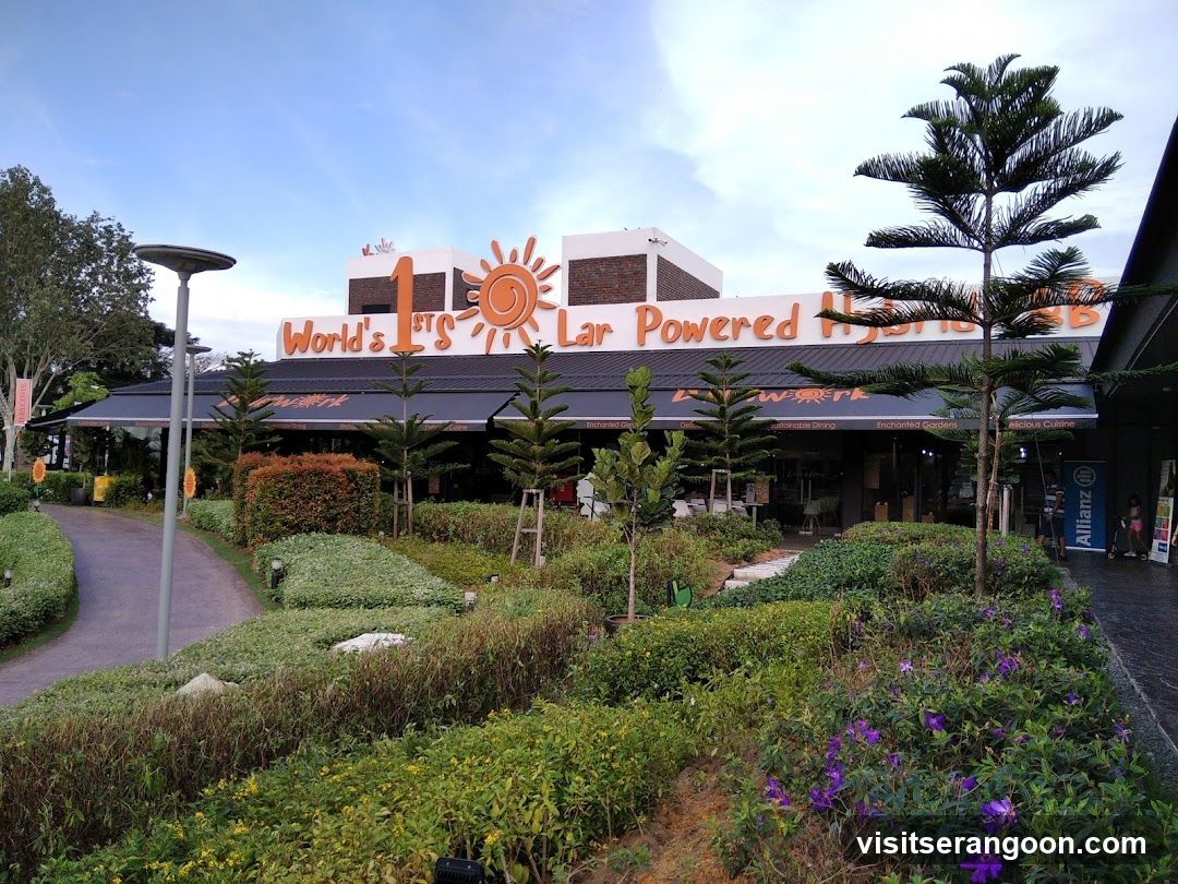 The exterior of a restaurant with a large sign that reads "World's 1st Solar Powered Hybrid". The building has a dark awning covering the entrance and windows. Landscaped gardens with various plants and flowers are in front of the building. A pathway leads to the entrance.