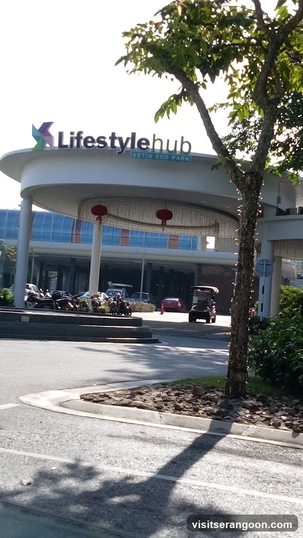 A wide shot of the entrance to Lifestyle Hub at Setia Eco Park. The sign "Lifestyle hub" is prominently displayed above the entrance, with "SETIA ECO PARK" written below it. The entrance features a curved overhang, and red lanterns hang from the ceiling. Several motorcycles are parked in front of the building, and a golf cart is visible in the background. The overall scene suggests a modern commercial or recreational complex.