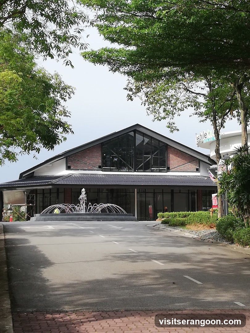 A wide shot of a modern building with a distinctive pitched roof and large glass windows. A circular fountain is in front of the building, with water spraying upwards. The building is surrounded by greenery and trees. A sign for 'Style Hub' is visible on a separate structure to the right.