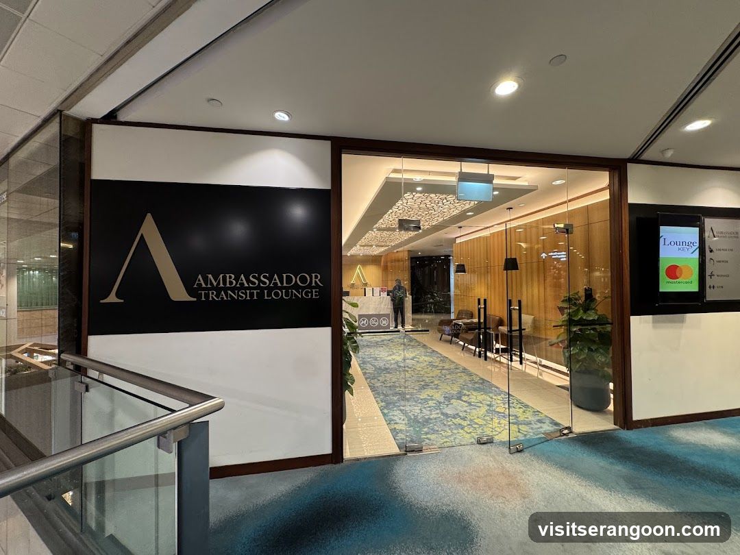 Photo of the Ambassador Transit Lounge storefront entrance, featuring a large sign with 'A' logo and text on black background. Glass doors open to interior lounge area with patterned carpeting, seating arrangement, and digital display board showing lounge key information.